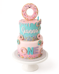 Two-tier donut themed birthday cake with pastel colours, sprinkles, drip icing and large donut topper on white cake stand
